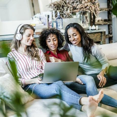 Trys laimingos moterys su nešiojamu kompiuteriu, sėdinčios ant sofos