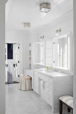 marmurinis vonios kambarys, marmuriniai stalviršiai, mažos marmurinės šešiakampės plytelės, baltos spintelės, baltas veidrodis, kosmetinė kėdutė ir taburetė